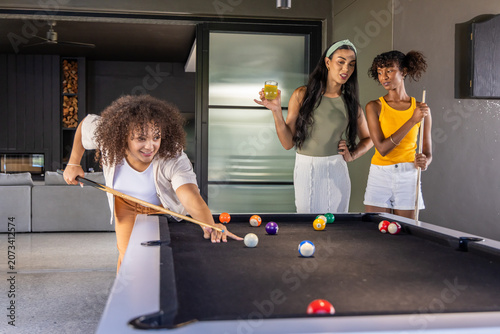 Diverse female friends playing pool in game room by black felt table with cues and balls