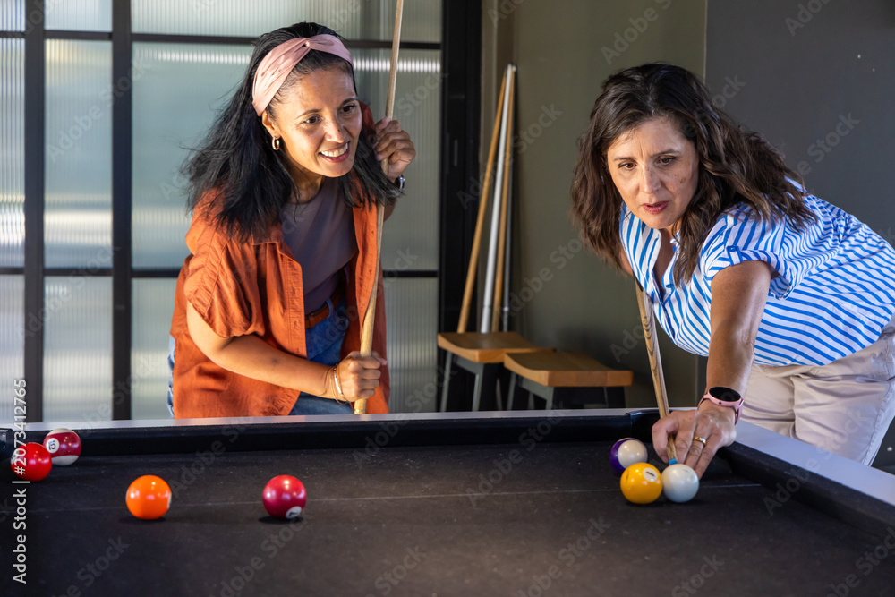 Naklejka premium Diverse female friends playing pool at black-felt table in rec room, holding cues and colored balls