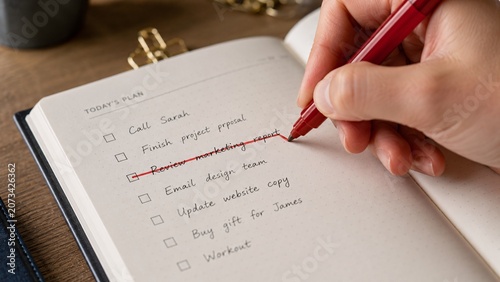 Close-up of a hand holding a red pen aggressively crossing out a finished task on a to-do list