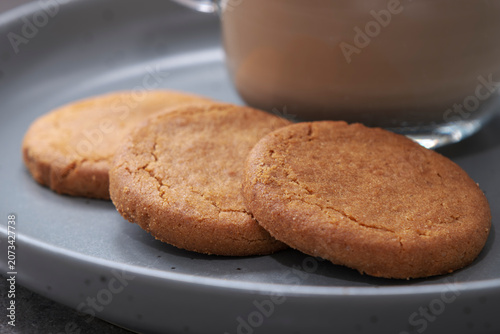 Golden cookies rest on a gray plate. Warm beverage sits softly in background. Simple, cozy snack scene. Ideal for cafes, breakfast, or comfort food themes. No people, no animals, no plants.