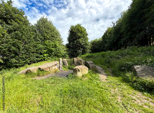 Lush green grass encircles an ancient stone circle in a serene forest clearing, while towering trees frame the view beneath a partly cloudy sky near Scaley Hills in Wibsey, UK