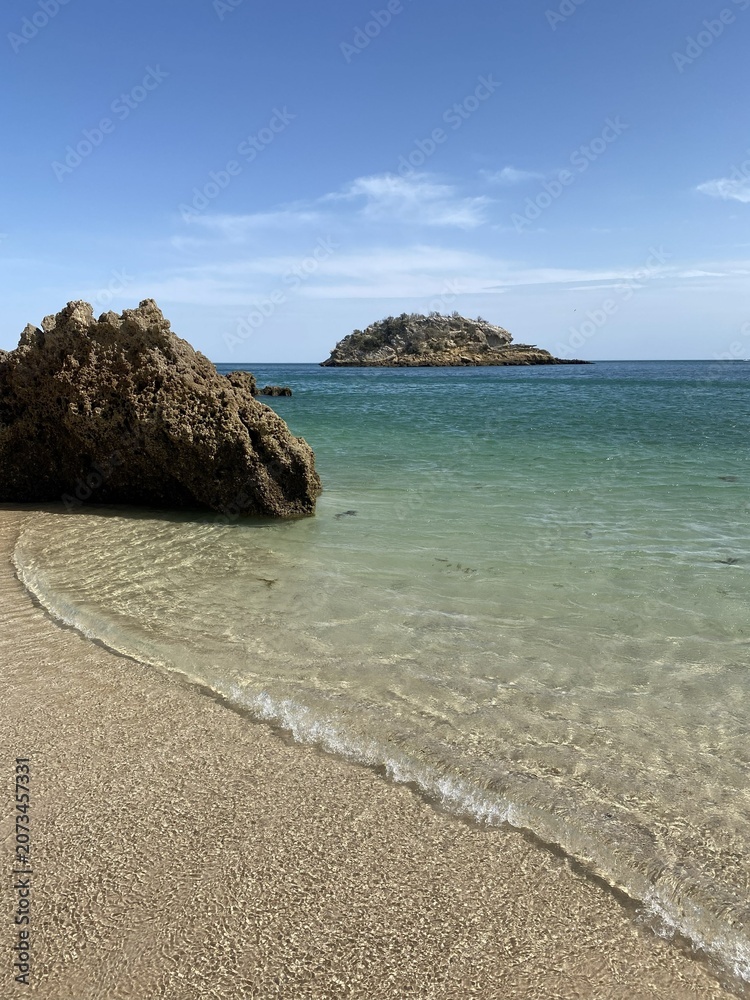 Obraz premium Praia de águas cristalinas com rochas e ilhéu ao fundo, areia clara e céu azul em cenário costeiro paradisíaco.