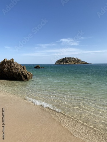 Quadro su tela Praia de areia dourada com águas cristalinas e ilhéu rochoso ao fundo sob céu azul, paisagem costeira tranquila de verão