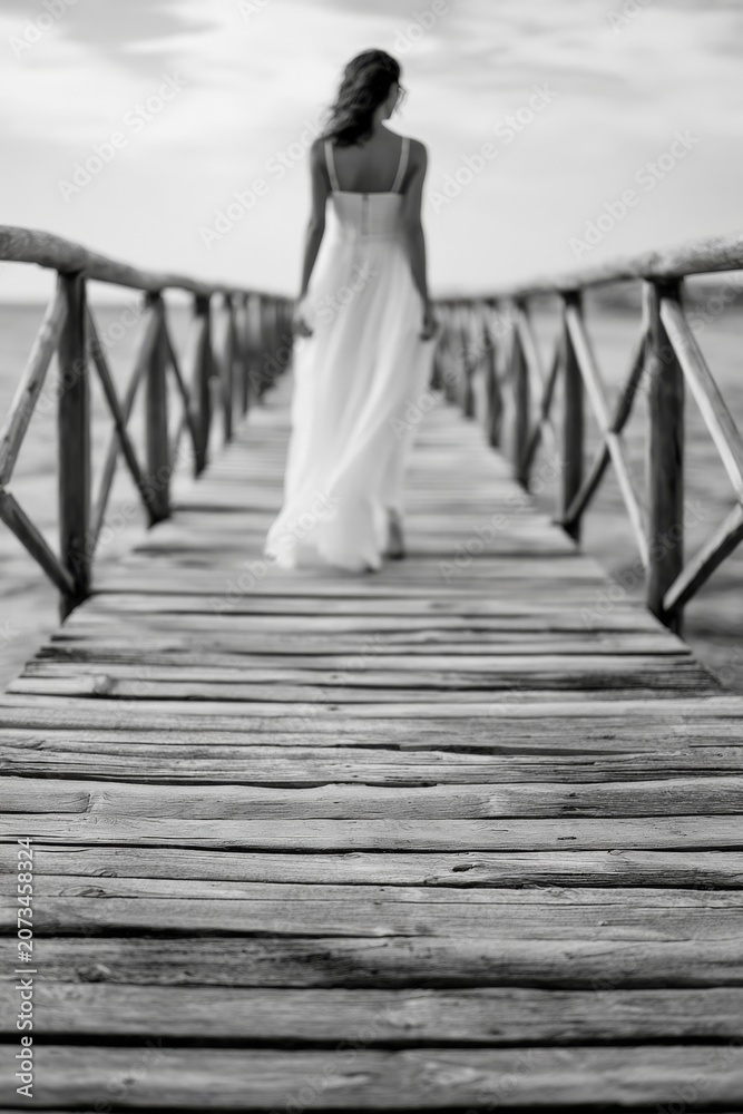 Fototapeta premium Woman in white dress walks on a pier while looking away into the distance at sunset