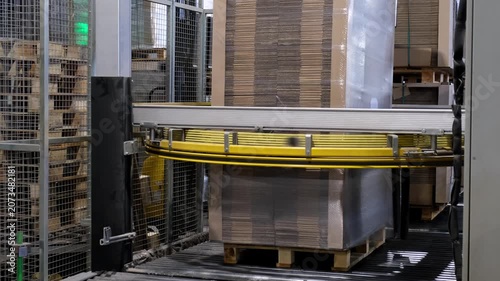 An automated factory packaging system wraps a tall stack of corrugated cardboard sheets on a wooden pallet using transparent plastic stretch film.