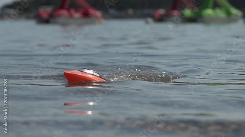 Slow motion video of small orange and black remote-controlled toy speedboat floating on the rippling blue water of a sea. The low-angle shot features a shallow depth of field, with colorful paddle
