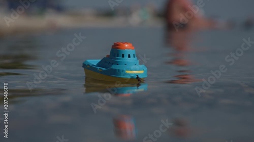 A small blue and yellow plastic toy boat floats on clear, calm water, casting a soft reflection. The low-angle shot captures the miniature vessel with a highly blurred background of distant watercraft