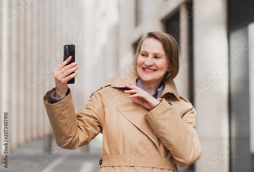 A mature smiling woman in a beige coat takes a selfie in an urban location on a spring day.