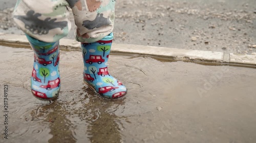 Close-up of a child wearing colorful rubber boots splashing through a rain puddle on a wet road. Water droplets fly through the air as small feet jump and play, capturing carefree childhood, outdoor