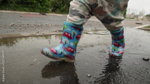 Close-up of a child wearing colorful rubber boots splashing through a rain puddle on a wet road. Water droplets fly through the air as small feet jump and play, capturing carefree childhood, outdoor