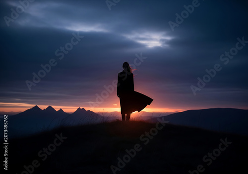 Woman in silhouette standing on hill during sunset with dramatic sky and mountain range