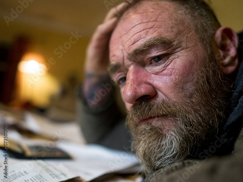 person sitting at a messy kitchen table hands rubbing their forehead in stress. Bills and papers spread out in front of them, a calculator nearby. . Expression of worry and exhaustion