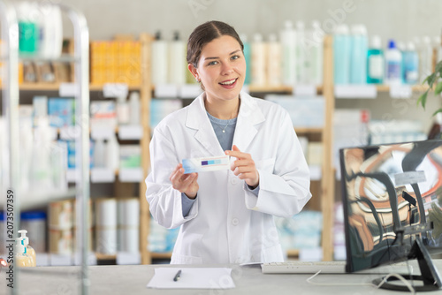 фотографія Smiling young female pharmacist in white coat standing at counter at drugstore a