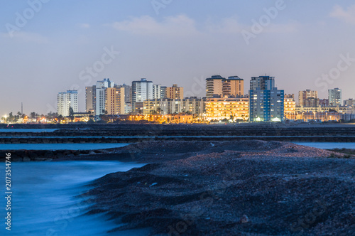 Long exposure of illuminated city skyline behind a rocky beach with silky water at night