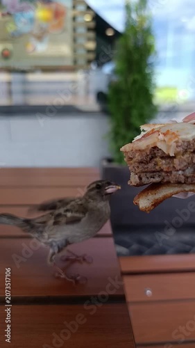 A sparrow is pecking at a piece of a burger on a wooden table outdoors.