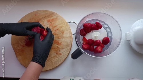 Hands in black gloves place strawberries into a blender for blending.