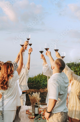 Group of friends holding wine glasses with red wine, toasting outdoors party in a vineyard. Winery, wine production, sunset time