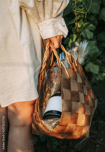 Young woman in white shirt carrying a wicker basket with a bottle of wine and wineglasses on outdoor in vineyard at sunset. Winery, wine production