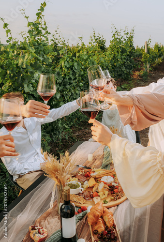Group of friends holding wine glasses with red wine, toasting outdoors party in a vineyard. Winery, wine production, sunset time