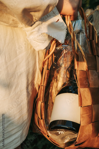 Young woman in white shirt carrying a wicker basket with a bottle of wine and wineglasses on outdoor in vineyard at sunset. Winery, wine production