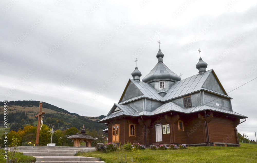 Obraz premium Traditional wooden Church of the Ascension in Bukovets village with dark timber walls and a tiered metal roof. It features three prominent domes topped with crosses against a cloudy, overcast sky