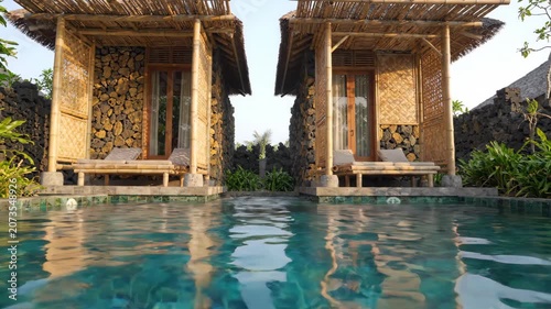 Bamboo cottage with stone walls and thatched roof beside clear blue pool and water in tropical sunny garden with lounge chairs