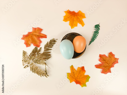 Top view flat lay of pastel colored eggs on a small plate decorated with autumn leaves on a white background.