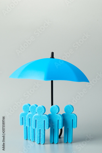A group of blue human figures standing together under a blue umbrella on a grey background. Group insurance, employee benefits, team protection, risk management concept. Copy space for text.
