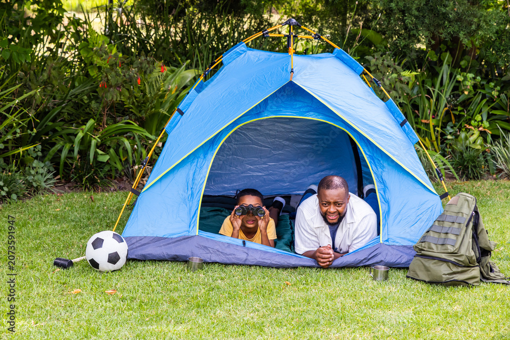 Fototapeta premium African American male father and child lying in backyard tent using binoculars beside backpack