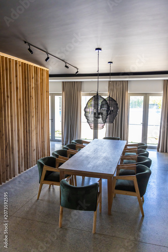 Long table anchoring dining area with ten green chairs, black pendant lamps, curtains, glass doors