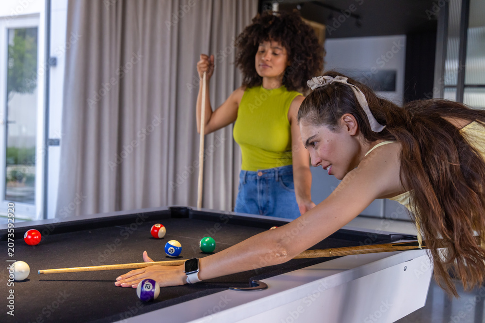 Fototapeta premium Diverse female friends leaning in game room black-felt pool table, aiming cue near striped balls