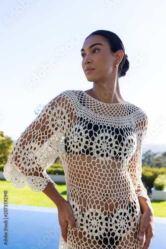 Adult African woman posing poolside wearing white crochet coverup over black swimsuit, gazing sea