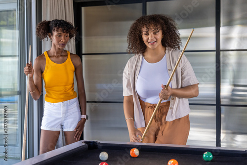 Diverse female friends wearing tank tops playing pool in modern lounge holding cue sticks and balls