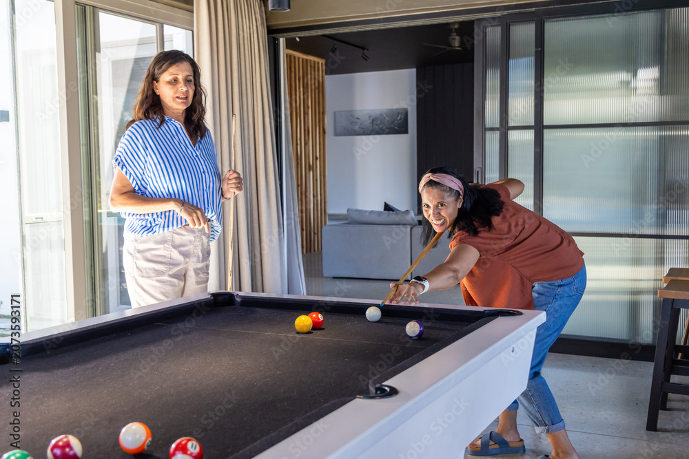 Naklejka premium Diverse women playing pool, lining up shots at home recreation area with black-felt table and cues