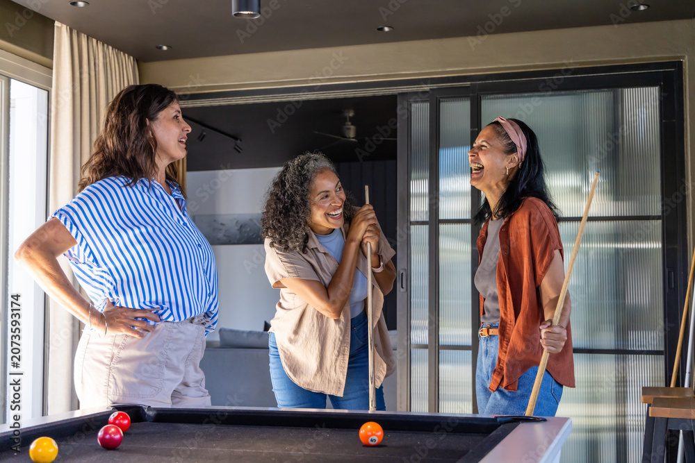 Naklejka premium Diverse female friends laughing, playing at pool table in home rec room, holding cue sticks
