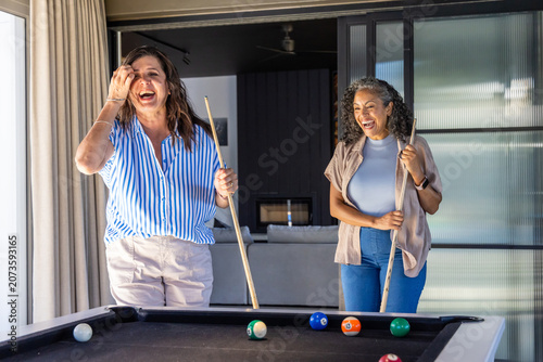 Diverse female friends laughing, playing pool with cues, billiard balls near sliding glass doors