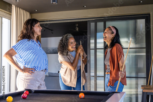 Diverse female friends laughing, playing at pool table in home rec room, holding cue sticks