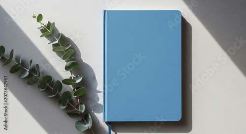 Elegant minimalist blue notebook journal with a vibrant green eucalyptus branch on a light background featuring harsh shadows creating a serene workspace scene for creativity