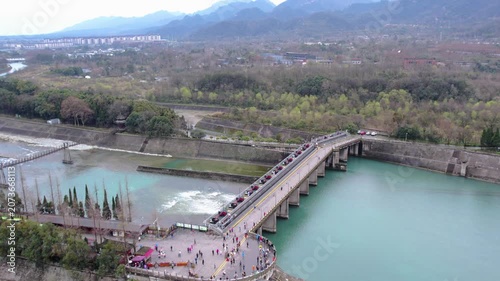 A historic hydraulic project site with a stone bridge spanning a turquoise river, surrounded by mountains and vegetation, with visitors on the bridge and square.
