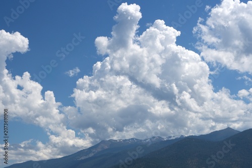 Billede på lærred Towering Clouds Over Snow‑Brushed Mountain Ridges — Dramatic cumulus formations rise above dark forested slopes and lingering patches of alpine snow