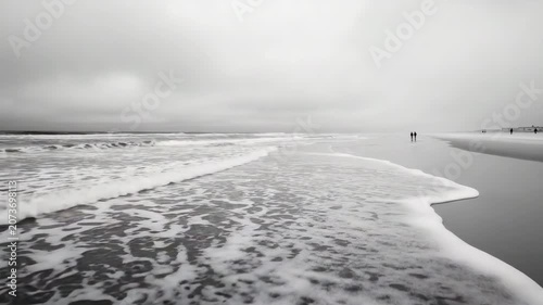 Two figures stroll on a quiet beach, where gentle waves lap against the shore. The cloudy sky creates a serene atmosphere as daylight fades into dusk.