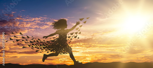 Silhouette of a young girl running and launching a flock of paper planes into the air against a bright sunset sky.