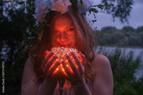 Young sexy woman wearing a floral wreath gently holds a glowing crystal lantern at twilight beside a quiet lake, creating an enchanting fairy tale scene filled with mystery, magic warm radiant light