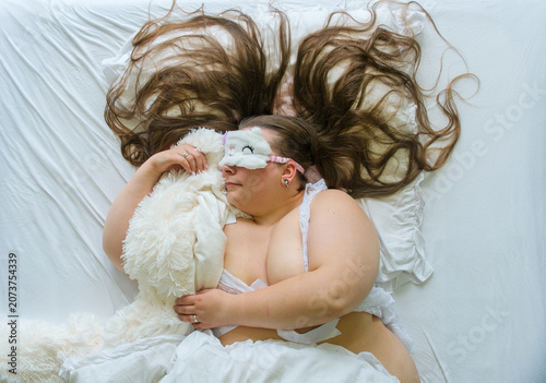 Curvy woman sleeping in white bed while wearing fluffy sleep mask and hugging plush toy creating a cozy bedtime scene with comfort relaxation and playful dreaming atmosphere.