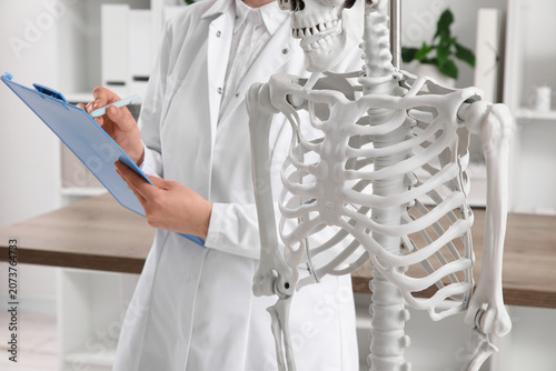 Female doctor writing on clipboard with human skeleton model in clinic