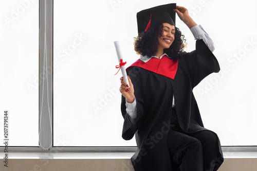 Happy African-American female graduate with diploma sitting near window
