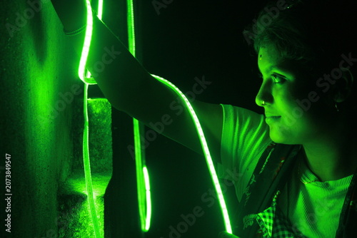 Creative portrait of a young Indian girl illuminated by vibrant green neon LED lights against a dark background. The dramatic lighting creates a futuristic atmosphere