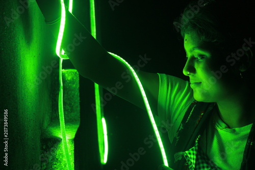 Creative portrait of a young Indian girl illuminated by vibrant green neon LED lights against a dark background. The dramatic lighting creates a futuristic atmosphere