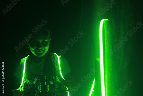 Creative portrait of a young Indian girl illuminated by vibrant green neon LED lights against a dark background. The dramatic lighting creates a futuristic atmosphere