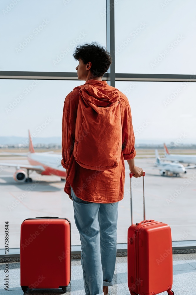 Obraz premium Young traveler standing at airport window with red luggage and backpack looking at planes for travel poster and background design